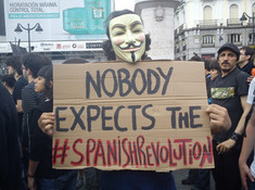Manifestante en la Puerta del Sol de Madrid el 17 de mayo. La frase es una referencia (détournement) al clásico de Monty Python Nobody expects the Spanish Inquisition!. Fuente: Wikimedia Commons.