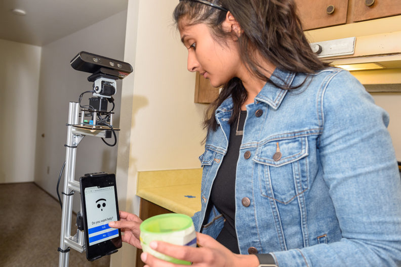 La estudiante Nisha Raghunath demuestra las interacciones entre un humano y un robot auxiliar en la Smart Apartment de la WSU, en Pullman. El robot detecta cuándo el sujeto comete un error en las tareas diarias y ofrece asistencia para ayudar a la persona a realizarlas. Imagen: Washington State University.